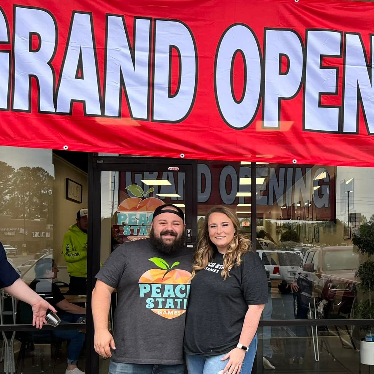 Two people standing in front of a grand opening banner at a business.